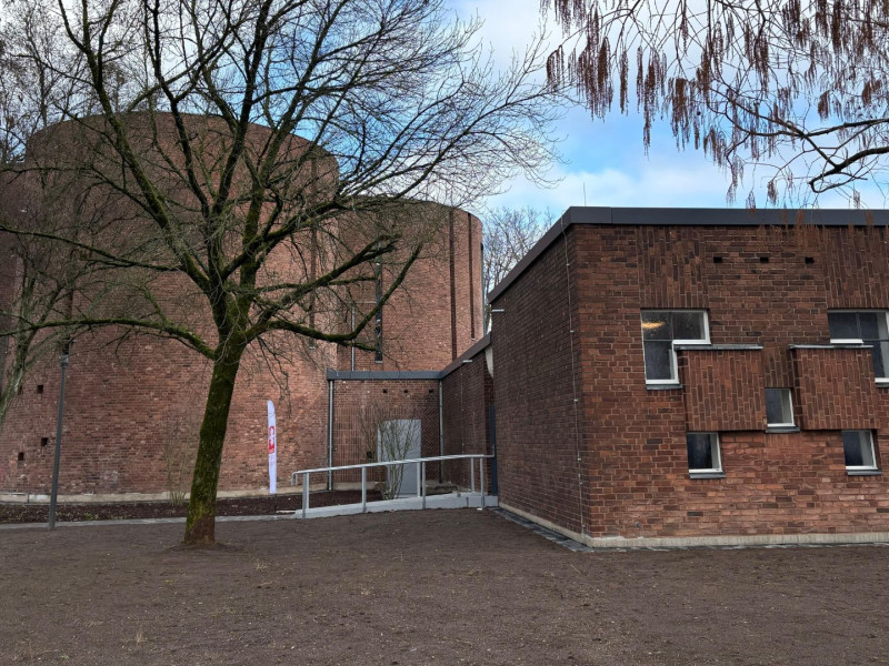 Außenansicht der ehemaligen Kirche St. Peter Morus in Leverkusen mit rotem Backsteinbau, rundem Kirchenschiff und modernem Anbau, umgeben von Bäumen.