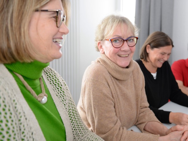 Es sind drei Frauen zu sehen, die an einer Besprechung teilnehmen. Eine von Ihnen lacht dabei direkt in die Kamera.