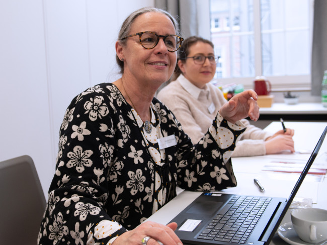 Zwei Mitarbeiterinnen des Hotels sitzen an einem Tisch, im Fokus ist eine der beiden Frauen, die vor einem Notebook sitzt. Beide Frauen scheinen aktuell an einer Besprechung teilzunehmen.
