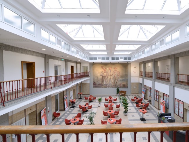 Innenansicht der Licht und Lohnhalle, es stehen mehrere gedeckte Tische im Foyer und Caritas-Plakate sind zu sehen. Das Foyer ist hell und man sieht, dass das Gebäude zweistöckig ist, zudem ein riesiges Gemälde mit Bergleuten.