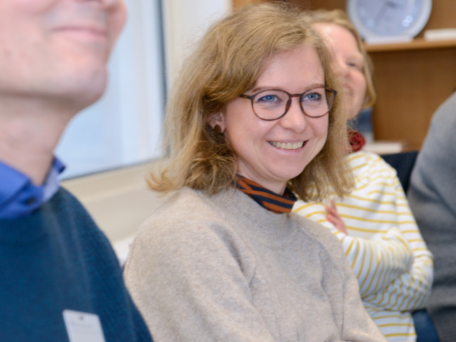 In einer Runde von Personen liegt der Fokus auf einer Frau, die in die Kamera lacht