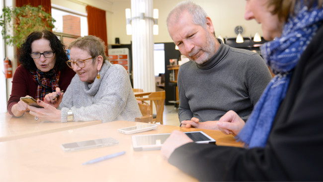 Menschen unterschiedlichen Alters sitzen im Vringstreff in der Kölner Südstadt an einem Tisch und benutzen Smartphones, während sie gemeinsam lachen und sich unterhalten.