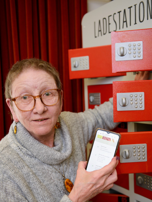 Eine Frau lächelt, während Sie mit ihrem Smartphone an den Stromladeschließfächern des Vringstreffs steht