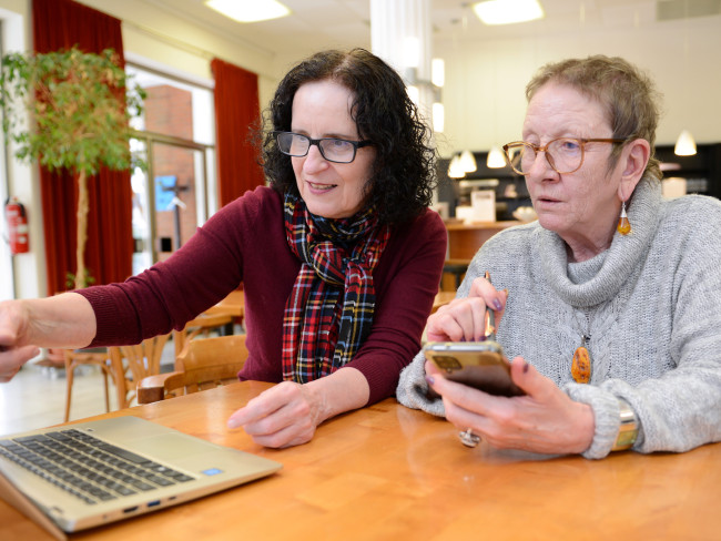 Zwei Frauen sitzen vor einem Notebook. Ein der Frauen zeigt auf das Notebook und erklärt der anderen Frau, die ein Smartphone in der Hand hält, etwas.