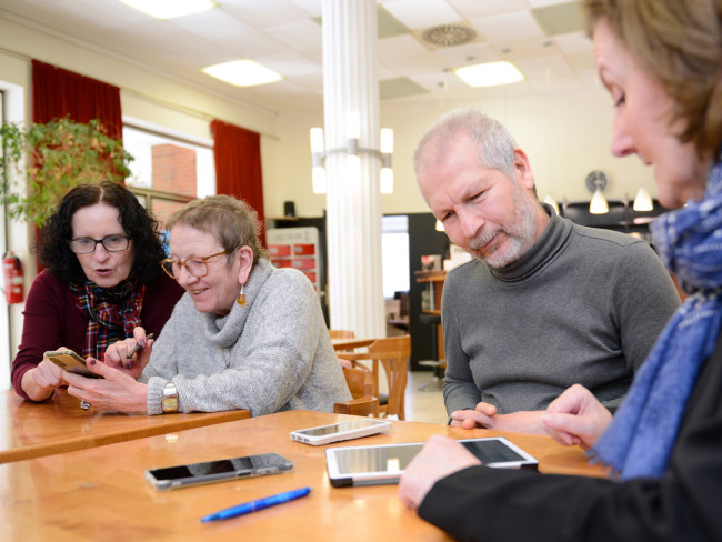 Mehrere Personen sitzen an einem Tisch und unterhalten sich, während verschiedene Smartphones auf dem Tisch liegen und ein Tablet. Einige der Personen haben auch ein Smartphone in der Hand, ihnen wird die Benutzung erklärt.