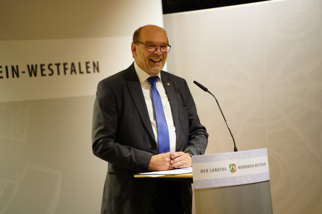 Lächelnder Redner steht am Pult bei Veranstaltung im Landtag Nordrhein-Westfalen.