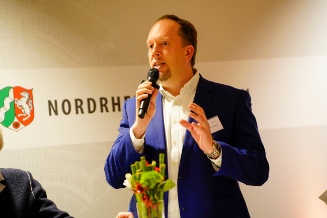 Mann im blauen Sakko spricht mit Mikrofon vor NRW-Wappen bei einer Veranstaltung.