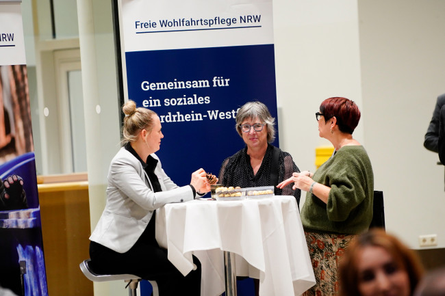 Drei Frauen im Gespräch an einem Stehtisch vor Roll-up der Freien Wohlfahrtspflege NRW.