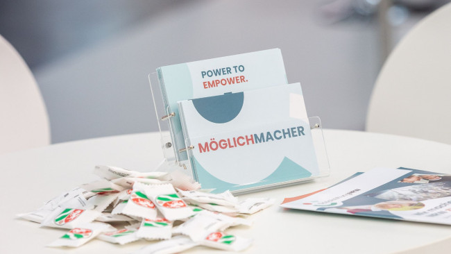 Infokarten mit Aufschrift „POWER TO EMPOWER“ und „MÖGLICHMACHER“ liegen auf weißem Tisch; daneben Giveaways und weitere REHACARE-Broschüren.