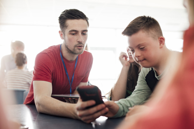 Mensch mit Einschränkungen schaut auf Smartphone und tippt. Daneben sitzt ein junger Mann und erklärt ihm etwas.